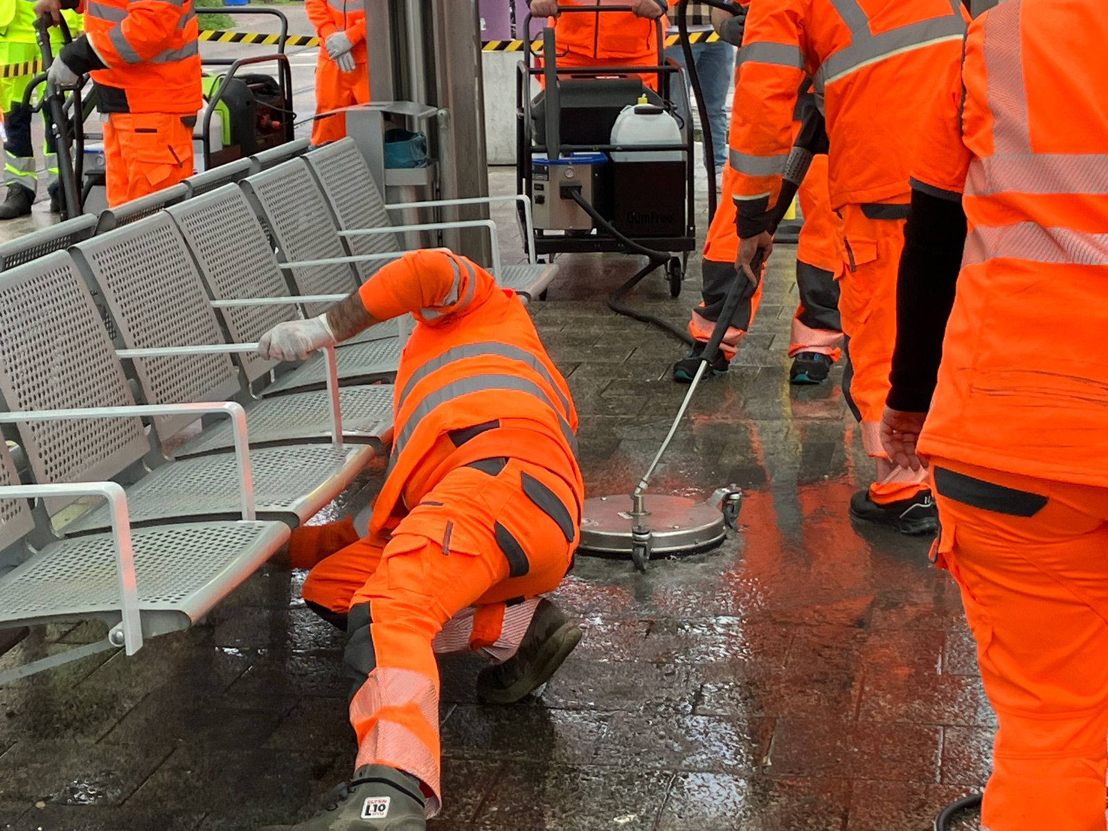 Mehrere Personen reinigen eine Haltestelle. Sie nutzen unterschiedliche Mittel und Geräte zum Putzen: einen Lappen, einen Hochdruckreiniger und ein spezielles Reinigungsgerät mit Düse. Die Personen tragen orangefarbene Warnkleidung. 