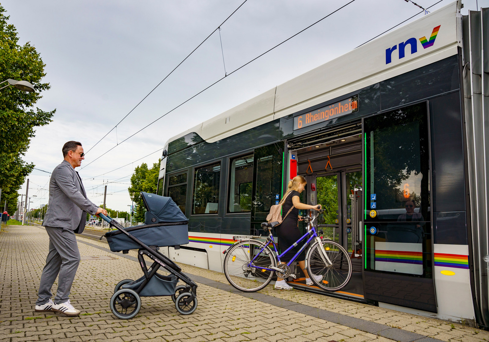 Ein Mann mit einem Kinderwagen und eine Frau mit einem Fahrrad steigen in eine Stadtbahn der rnv vom Typ Rhein-Neckar-Tram (RNT) ein.