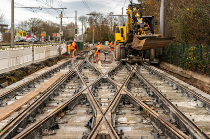 Gleisbauarbeiten an einer Straßenbahnkreuzung mit mehreren spitz zulaufenden Schienenprofilen im Zentrum, an denen Personen in Warnkleidung arbeiten. Ein gelbes Baustellenfahrzeug steht seitlich an den Gleisen.