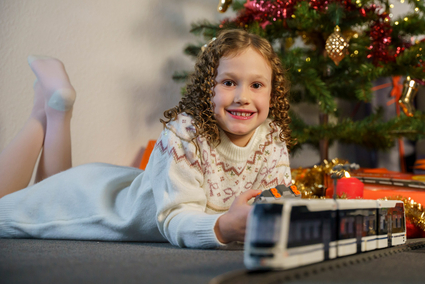 Ein Mädchen mit dunkelblonden Korkenzieher-Locken in einem Strickkleid liegt vor einem festlich geschmückten Weihnachtsbaum und lächelt. Im Vordergrund fährt das Klemmbaustein-Modell einer Straßenbahn vom Typ Rhein-Neckar-Tram (RNT).