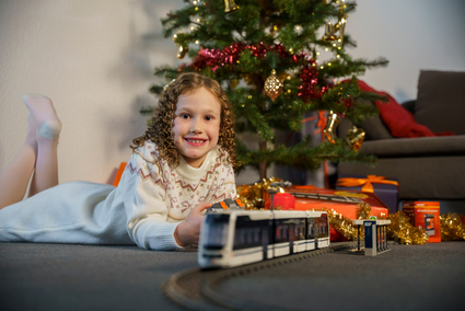 Ein Mädchen mit dunkelblonden Korkenzieher-Locken in einem Strickkleid liegt vor einem festlich geschmückten Weihnachtsbaum und lächelt. Im Vordergrund fährt das Klemmbaustein-Modell einer Straßenbahn vom Typ Rhein-Neckar-Tram (RNT).