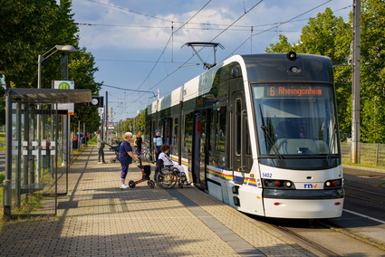 Eine Stadtbahn der rnv vom Typ Rhein-Neckar-Tram (RNT) steht mit geöffneten Türen an einer Haltestelle. Es steigen Menschen in die Bahn: Eine Frau im Rollstuhl, eine Frau mit Rollator, ein Mann ohne Beeinträchtigung, ein Mann mit Kinderwagen und eine Frau mit Fahrrad.