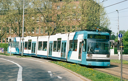 Eine türkisene Straßenbahn fährt um eine Kurve. Im Hintergrund sieht man ein altes, prächtiges Gebäude und viele große Bäume.