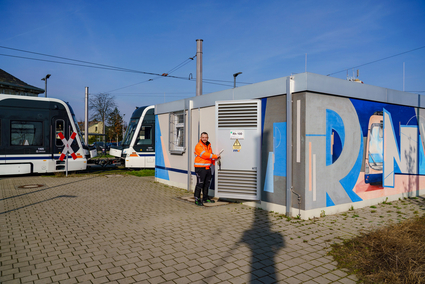 Ein Mitarbeiter der rnv in oranger Sicherheitskleidung steht in der geöffneten Tür eines Gleichrichterunterwerks und blick in die Kamera. Im Hintergrund fährt eine Straßenbahn vom Typ Rhein-Neckar-Tram vorbei.
