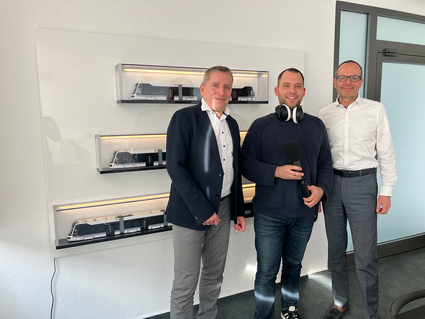 Die Geschäftsführer der Rhein-Neckar-Verkehr GmbH (rnv), Martin in der Beek und Christian Volz stehen links und rechts neben Moderator Jens Schneider in einem Besprechungszimmer. An der Wand hinter ihnen sind drei Modelle der Rhein-Neckar-Tram in den drei Längenausführungen (30, 40 und 60 Meter) auf Regalbrettchen ausgestellt.