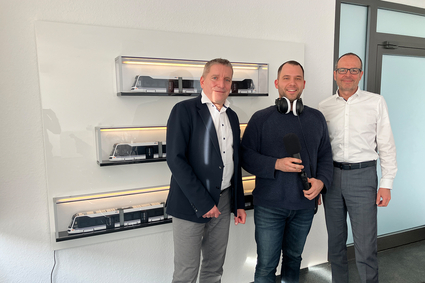 Die Geschäftsführer der Rhein-Neckar-Verkehr GmbH (rnv), Martin in der Beek und Christian Volz stehen links und rechts neben Moderator Jens Schneider in einem Besprechungszimmer. An der Wand hinter ihnen sind drei Modelle der Rhein-Neckar-Tram in den drei Längenausführungen (30, 40 und 60 Meter) auf Regalbrettchen ausgestellt.