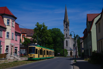 Eine beige-grüne Straßenbahn fährt auf Rasengleis durch Schöneiche. Im Hintergrund stehen große, alte Bäume und ein markanter Kirchturm.