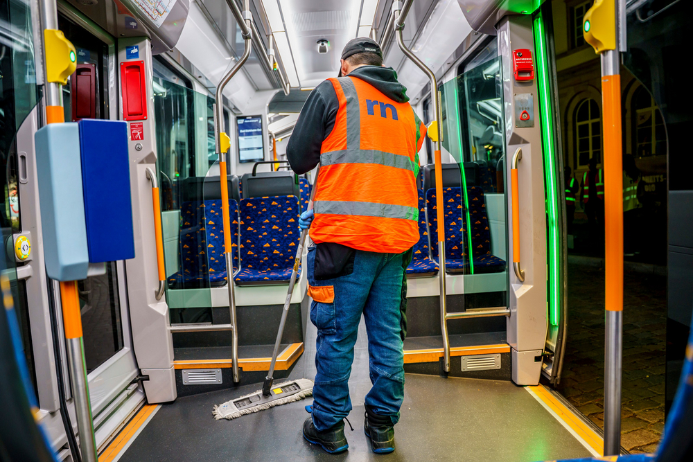 Ein Mitarbeiter mit orangefarbener rnv-Warnweste steht in einer Straßenbahn vom Typ RNT. In der Hand hält er einen Wischer, um das Fahrzeug zu reinigen.