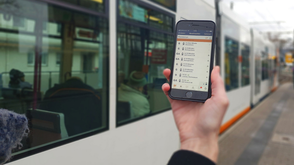 Hand hält ein Smartphone an einer Stadtbahnhaltestelle