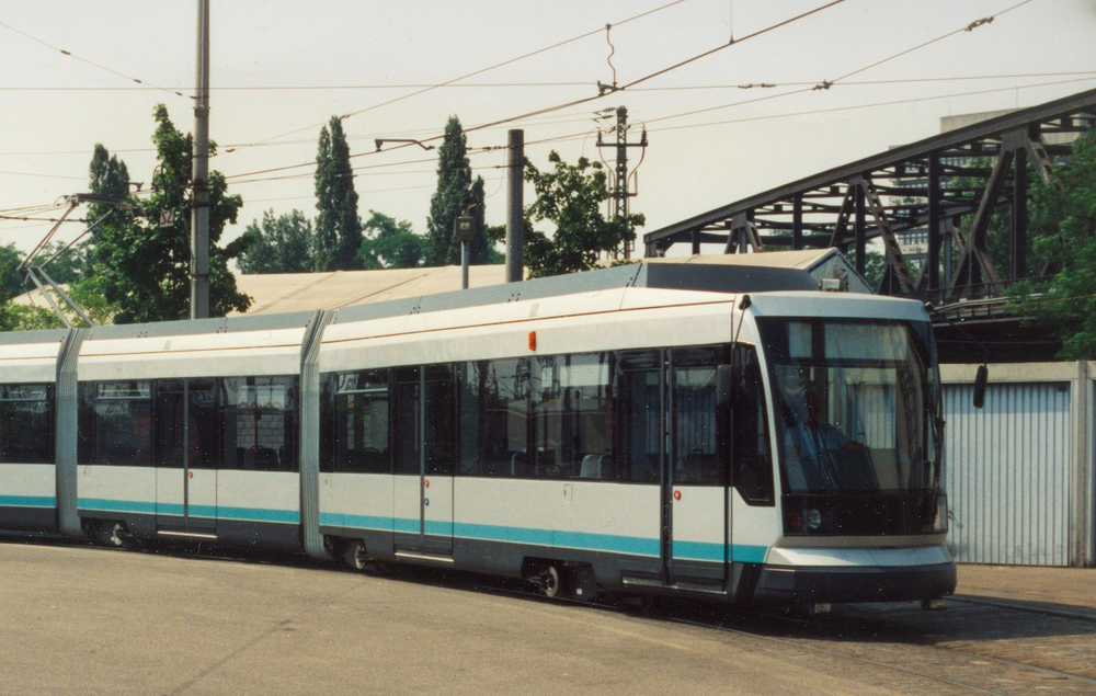 Ein metallic-farbener Straßenbahn-Prototyp steht auf dem Betriebshof. Im Hintergrund ist eine alte Stahlbrücke zu sehen.